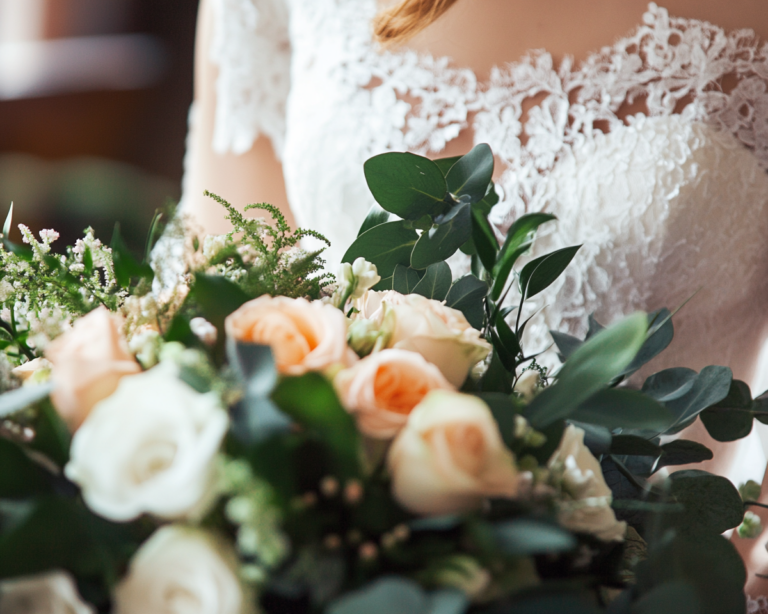 La photo montre un bouquet de mariage élégant avec des roses blanches et pêche, entourées de feuillage vert, porté par une personne en robe de mariée en dentelle délicate. L'image est lumineuse et douce, capturant l'essence romantique et raffinée d'une cérémonie de mariage.