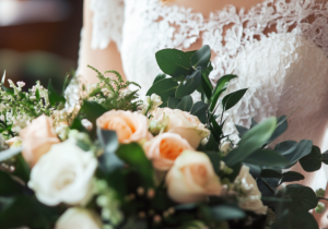 La photo montre un bouquet de mariage élégant avec des roses blanches et pêche, entourées de feuillage vert, porté par une personne en robe de mariée en dentelle délicate. L'image est lumineuse et douce, capturant l'essence romantique et raffinée d'une cérémonie de mariage.